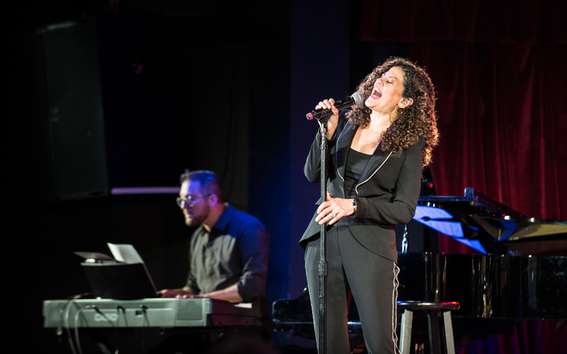Karla Harris singing into a microphone on stage as a man plays piano