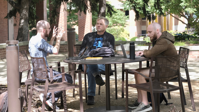 Philosophy master's student Logan Bohlinger talks with Professor Eric Wiland and student Daniel Grasso while seated at a table in the Quad