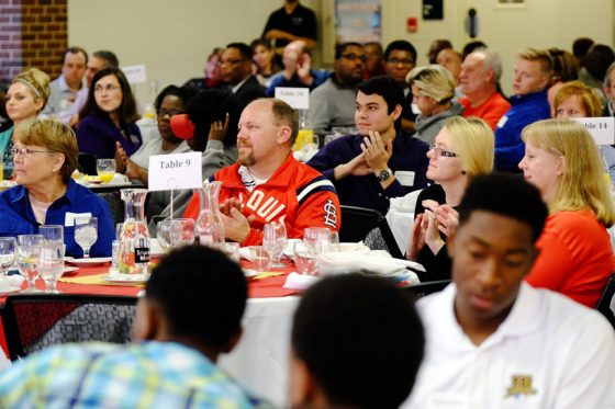 A diverse group of people sitting around round tables in a crowded room applauds.