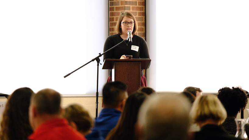 UMSL student Kathryn Loucks looks out over a crowd of people while standing at a lectern on stage and speaking into a microphone.