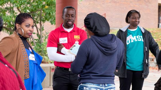 UMSL students Cynthia Davis, Tim Lewis and Kimberly Brown stand under and overhang and observe as a woman with a hat fills out a survey after voting.