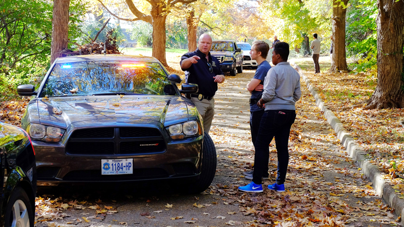 Trooper Scott Miller stands next to a parked Missouri State Highway Patrol car and points as he talks to criminology and criminal justice students Sherman Brawner and Marcella Hamilton on a shaded street.