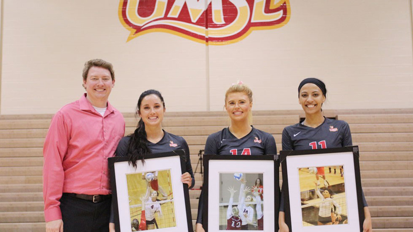 UMSL coach Ryan Young poses with senior volleyball players Samantha Lewis, Gaylynn Jones and Lexi Rogers. The players are all holding framed pictures of them in action on the court.