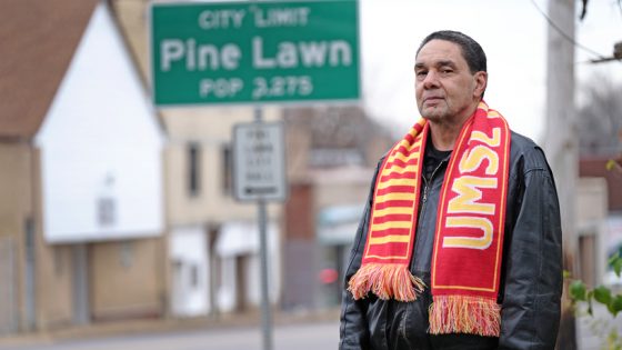 Elwyn Walls standing in front of the Pine Lawn city limit sign while wearing an UMSL scarf