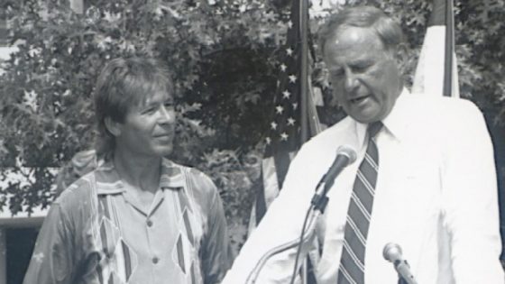 Robert R. Hermann speaks at a microphone with John Denver by his side