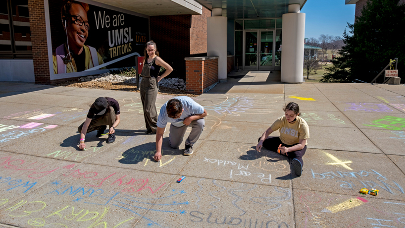 Three students squatting decorate the quad with chalk while one looks on