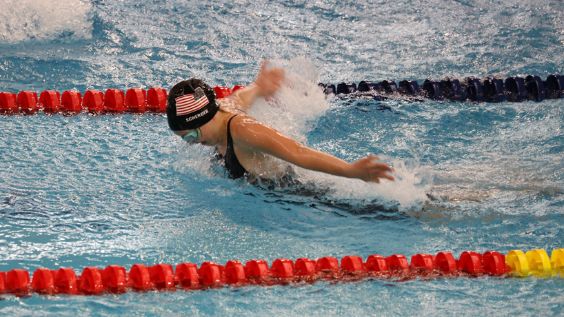 Maggie Scherder, white woman in blue swim cap with American flag and goggles breaches the surface of the a pool while performing a breaststroke.