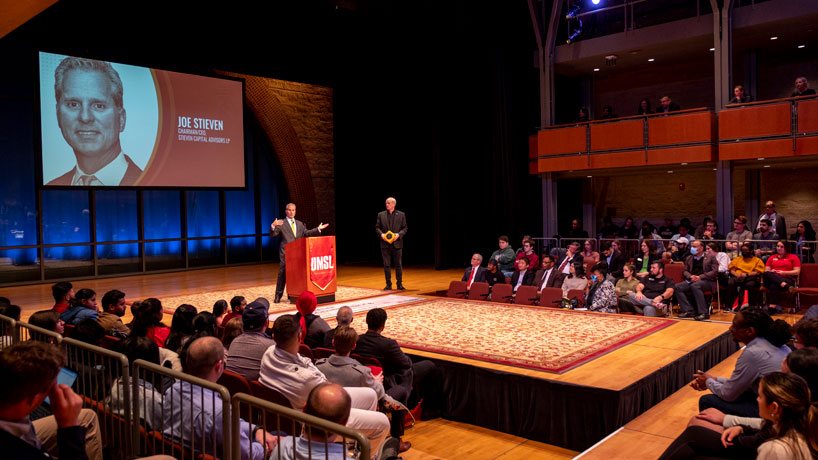 A man stands at podium on a stage with an audience while another man stands to the side.