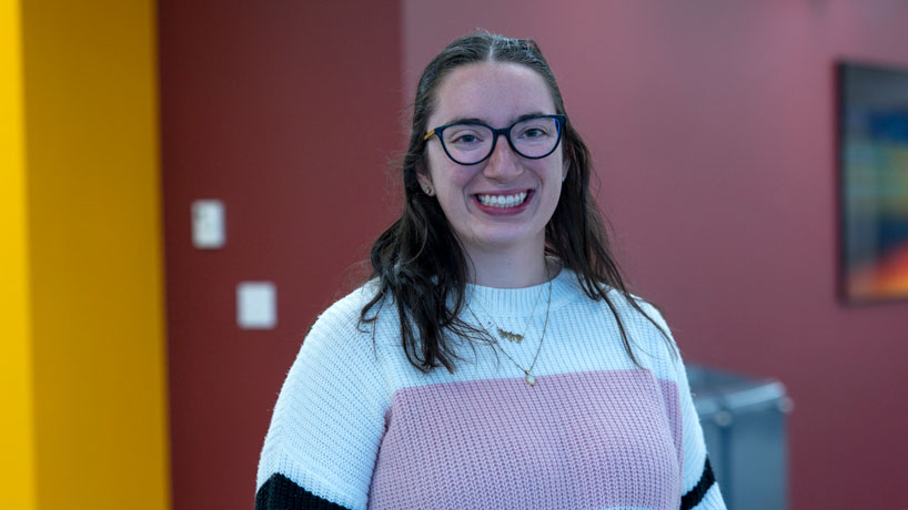 Young white woman wearing glasses and a sweater looks straight ahead, smiling.