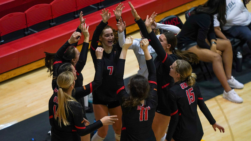 UMSL volleyball players huddle together and celebrate with their arms in the air