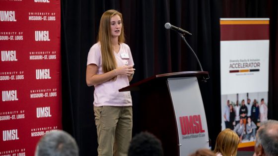 Young, white woman with long brown hair stands at a podium speaking to a crowd.