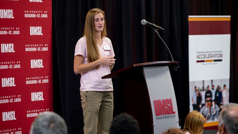 Young, white woman with long brown hair stands at a podium speaking to a crowd. 