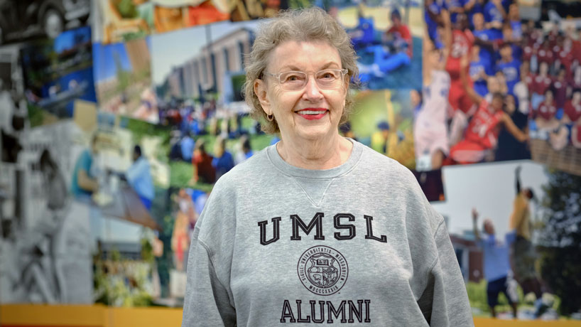 Older white woman wearing glasses and an UMSL sweatshirt, stands, smiling against a colorful collage of photos.