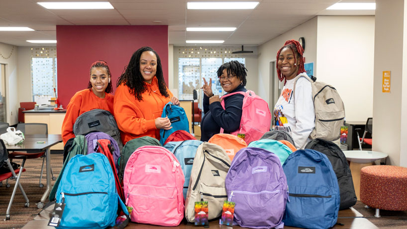 Four Black, female students who are smiling, stand behind a table that has different colored backpacks.