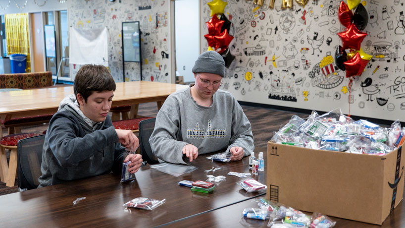 Two white young women sit at a table assembling gift bags.