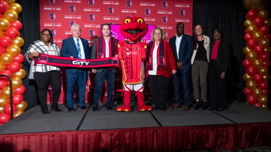 A group of UMSL staff and faculty members stand next to Louie, the Tritons red salamander mascot, in front of red CITY SC backdrop