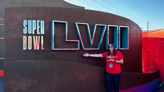 Kalyn Ohrt, white woman in red shirt an sunglasses, gestures in front of Super Bowl sign