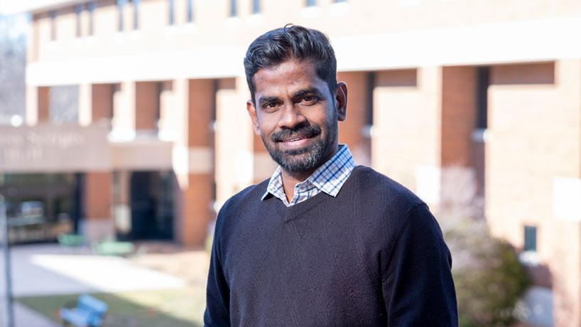 An Indian man stands outside in front of a building on a university campus.