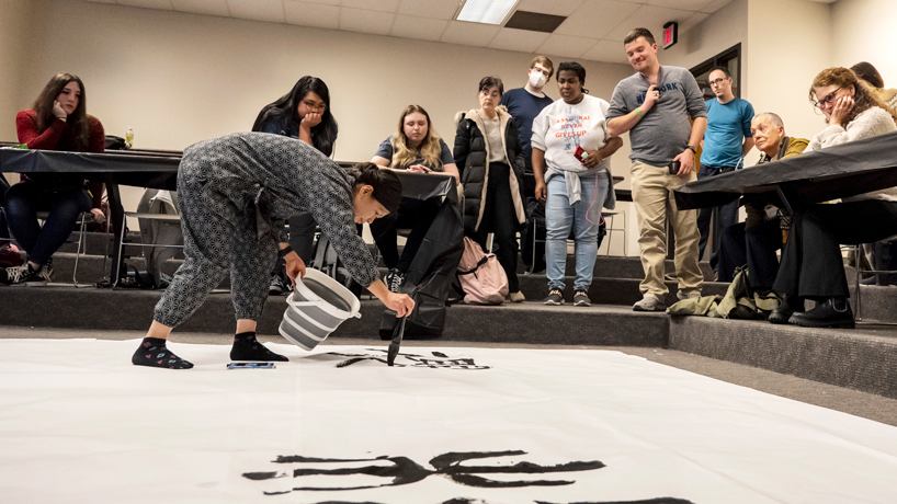 Kaori Ishijima stands on calligraphy paper and uses a brush to write the letter for sakura in Japanese while students watch