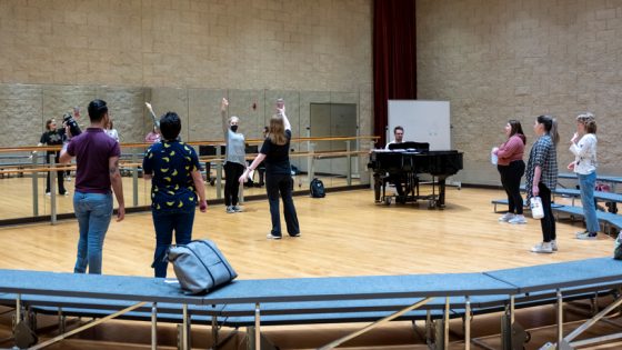 Karen Kanakis, the Interim Director of Opera Theater, white woman in mask and gray shirt and black pants, leads several students in vocal exercises in studio with wood floor and mirror