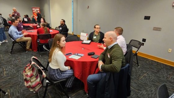Young woman is talking with a recruiter, sitting at a table in a room full of people also sitting at tables at a networking event.