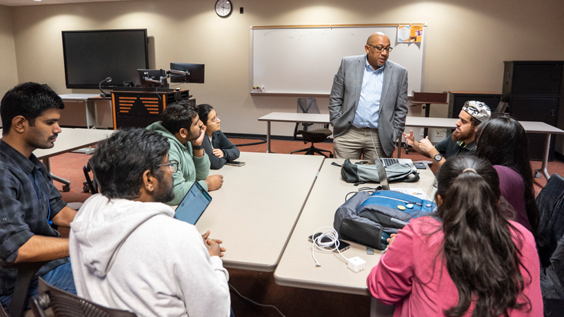 Assistant Teaching Professor Damon Walker stands as he listens to a student in his IT Project Management class while other students listen