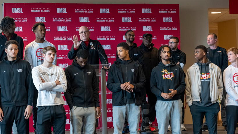 Coach Bob Sundvold speaks on stage while surrounded by his players at a men's basketball pep rally 