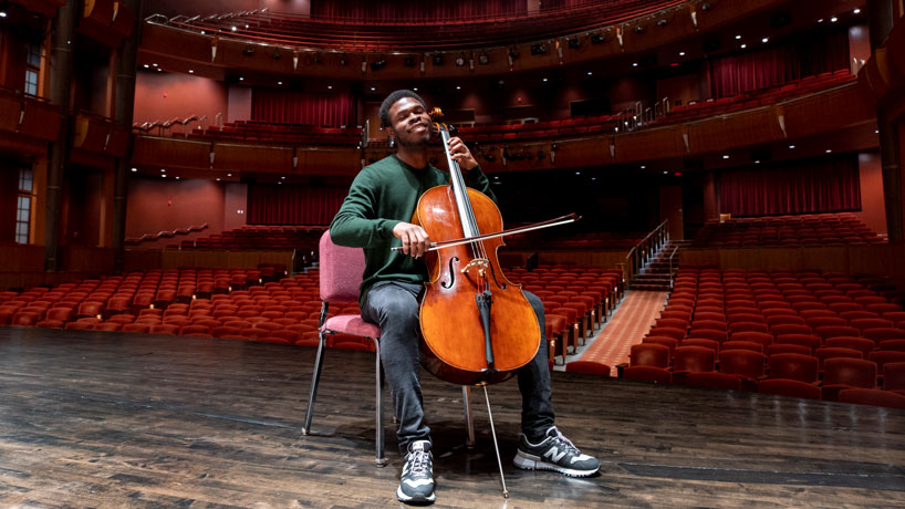 Young black male sits on chair playing the cello on stage with his back to the empty auditorium.