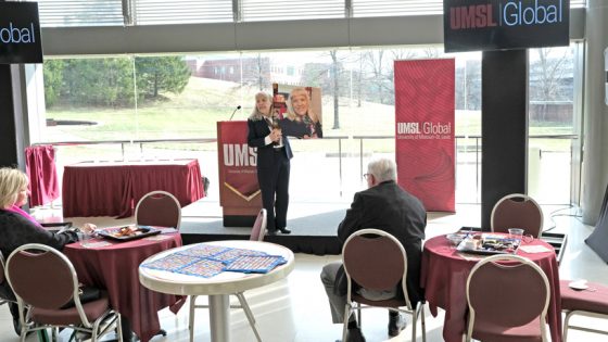Professor Anne Fish holds up a life-size portait of herself made in cross stitch at the UMSL Global Faculty Reception