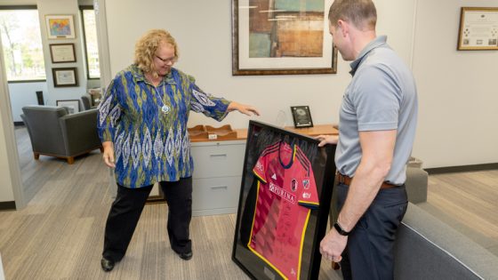 Kristin Sobolik, white woman in blue top and black pants, looks at framed soccer jersey as white man in a gray polo looks on