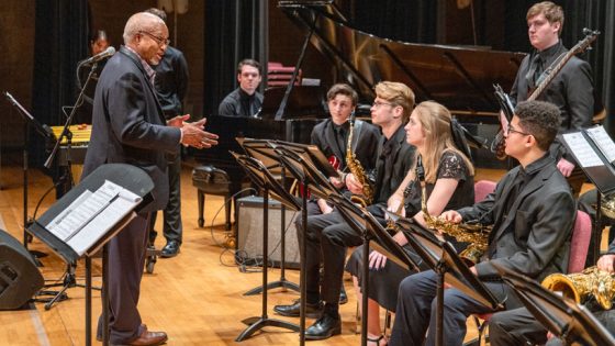 A black male music clinician addresses jazz music students in a rehearsal on stage in an auditorium.