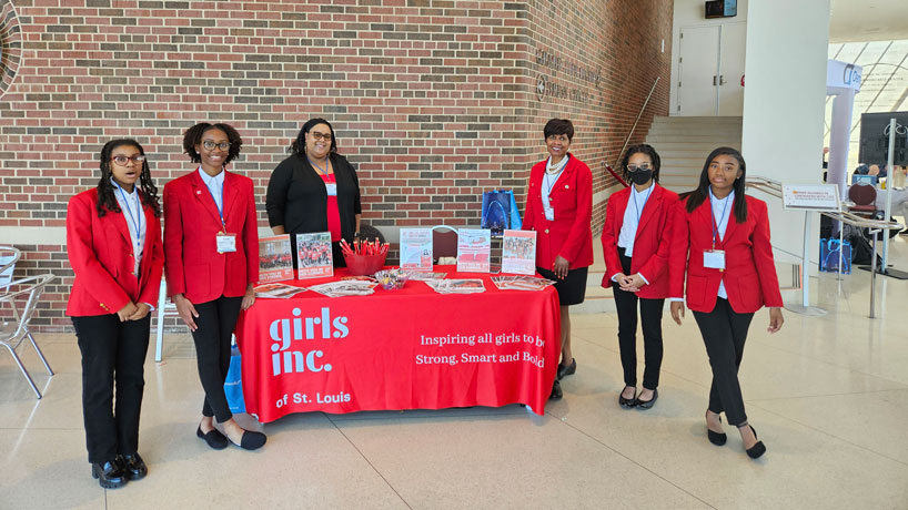 Several black high school girls and two black women stand at the Girls Inc. table posing and smiling.