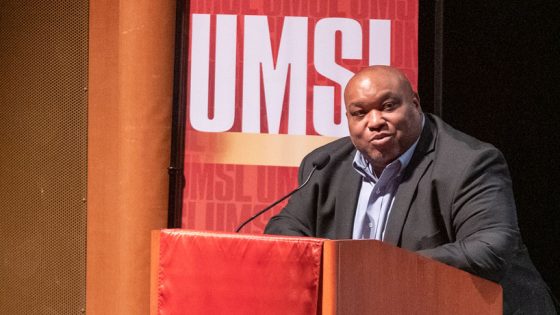 Black man stands at podium in front of an UMSL sign addressing students in an auditorium.