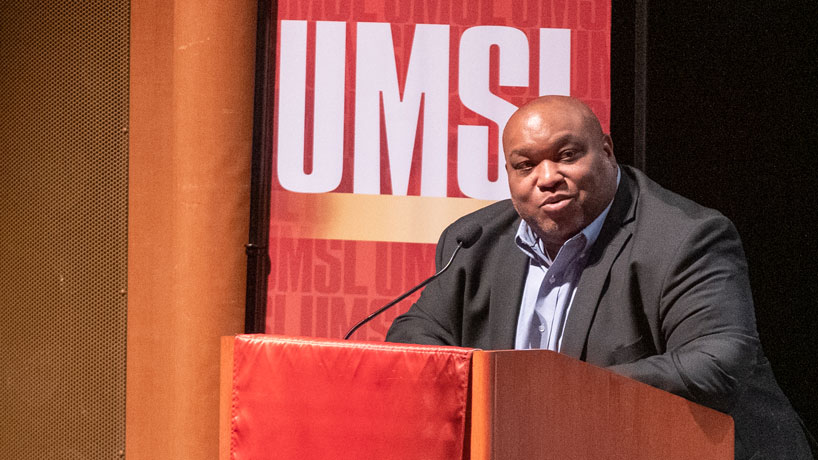 Black man stands at podium in front of an UMSL sign addressing students in an auditorium.