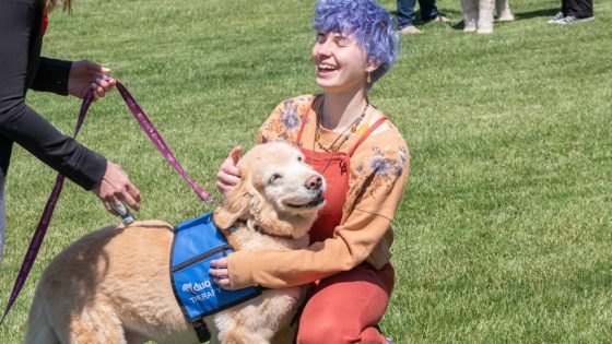 Young white female student with purple hair plays with dog on grass