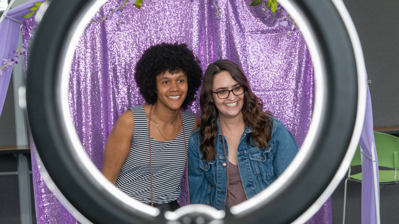 Brown woman and her girlfriend pose for a photo against a lavender backdrop.