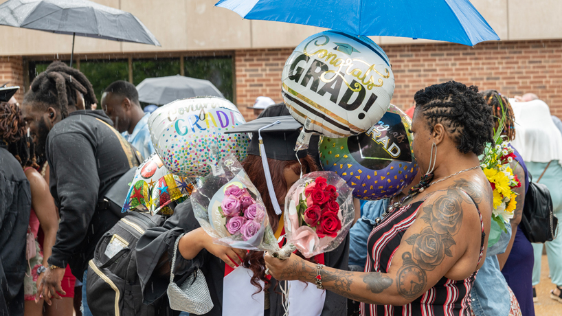 Friends and family, black women, cover a graduate, black woman in black cap and gown, with flowers and balloons. 