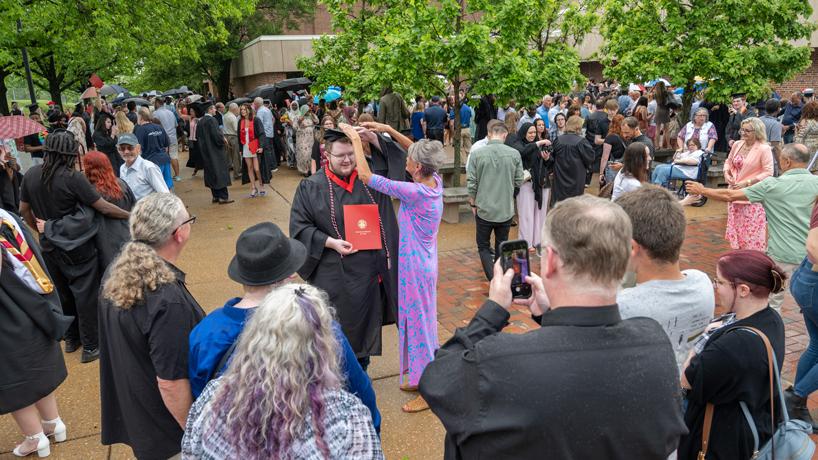 UMSL graduate, white man in black cap and gown, stands while white woman in pink and blue dress adjusts his cap