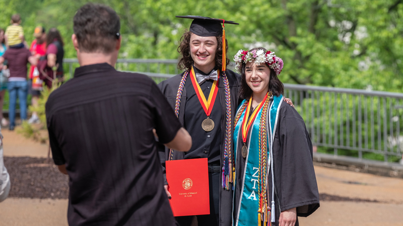 Drew Ryherd, white man with long dark hair in black cap and gown, and Sydney Stark, white woman in flower crown and black gown, pose for photos