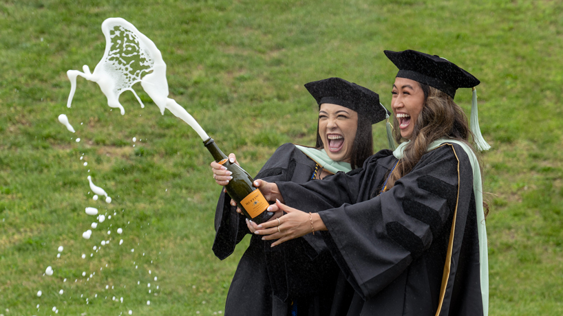Tiffany Lee and Chanell Nguyen, two asian women in black cap and gowns, pop a bottle of bubbly
