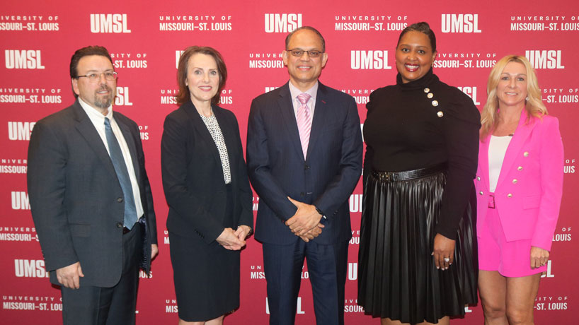 Several, diverse UMSL alumni stand infront of an UMSL backdrop smiling