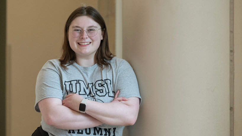 Emily Sigmund leans again a wall, smiling with her arms crossed while wearing an UMSL alumni T-shirt