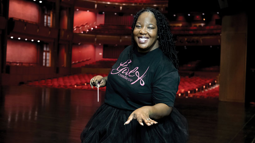 Black woman wearing all black, holding a conductor wand, stands on stage at the Touhill theater with a red background of theater seats behind her