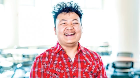Cin Lam Mung, asian man with frizzy hair in red plaid western shirt, smiles in classroom