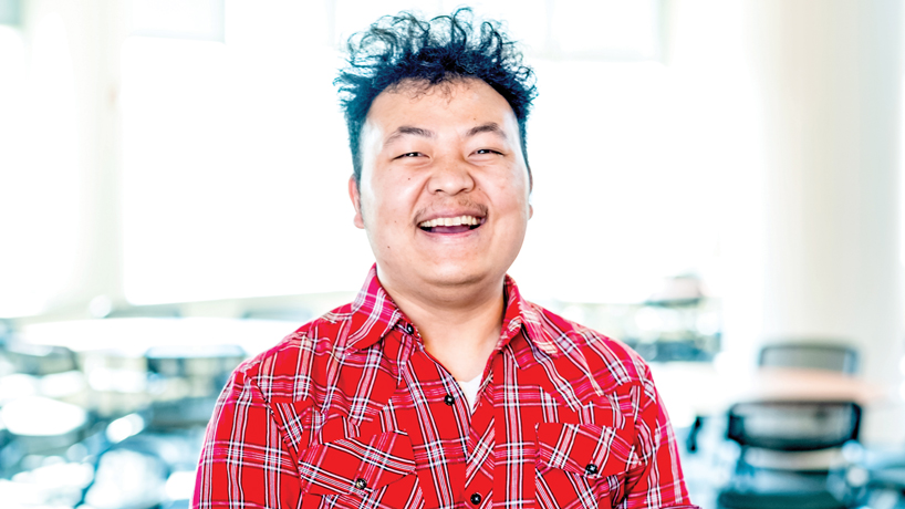 Cin Lam Mung, asian man with frizzy hair in red plaid western shirt, smiles in classroom