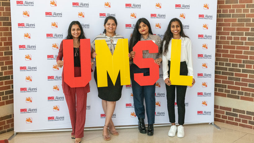 Four UMSL graduates stand together holding red and gold letters that spell out UMSL