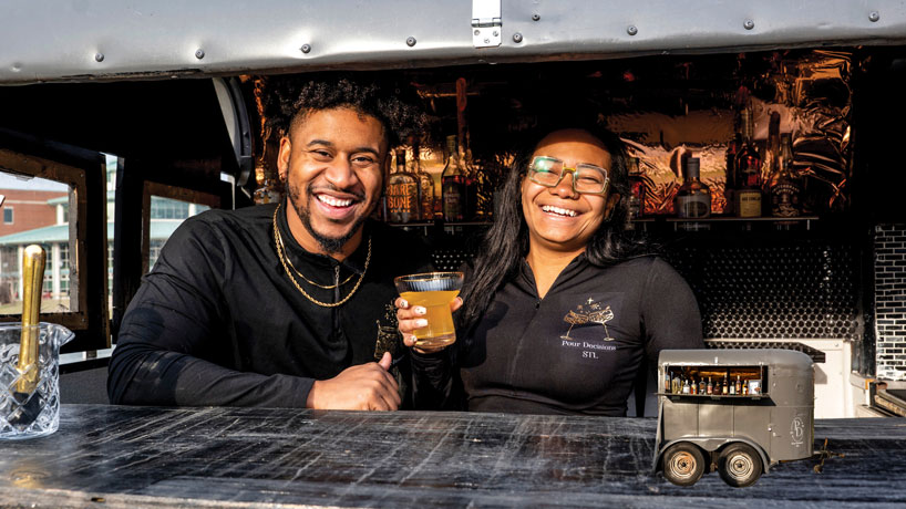A black couple dressed in black are in a food truck smiling. The woman is holding a drink.