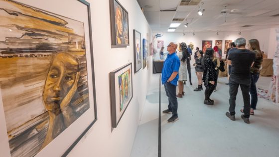 a water water color painting with earthtones and black is displayed on a white wall while people look at other paintings and talk to each other in the background