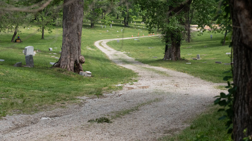 A gravel roadway runs through a cemetery with grass and trees and some visible headstones.