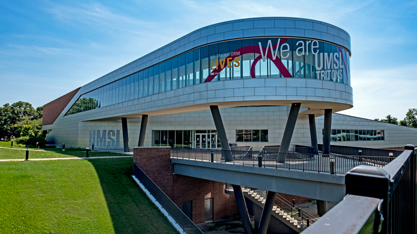 the Recreation and Wellness Center, a sleek metallic building with some brick elements, is pictured looking toward the entrance with a blue sky and clouds behind it